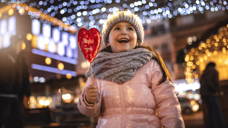 Image showing a child at a giving season event like a Winter Carnival. She is dressed in warm clothing, holding a treat, and smiling while looking up at holiday lights.